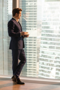 Pensive businessman in stylish business suit drinking coffee while looking on city view through large window in office, luxury apartments or hotel room. Relaxed successful CEO thinking about future