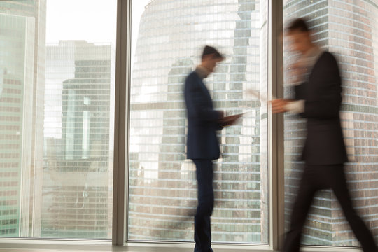 Image of blurry silhouettes of office workers hurrying in business past large window with city view in modern office. Fast and tense rhythm of modern business life, busy businessmen, economical grow