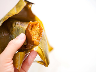 Side view of peeled untied bitten Thai Chinese style brown steamed minced nut stuffed dough pyramid dessert in yellow green banana leaf, held by left lady woman hand, white background