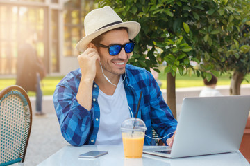 Closeup portrait of young handsome Caucasian man in street cafe in process of joyful online communication via laptop and earphones, listening to positive news and jokes, looking interested and happy