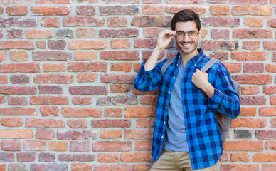Fototapeta na wymiar Horizontal image of sporty European man pictured with red brick wall behind ready for discovering new places with backpack on his back and fashionable black-rimmed spectacles lit with daylight