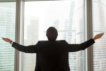 Successful businessman in dress suit standing in office in front of sunshine lit window with arms wide open. Welcoming new successful deals, business opportunities and bright future concept. Back view