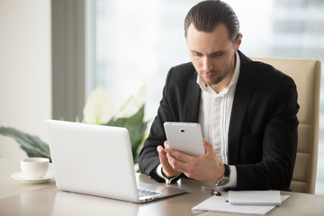 Smart young serious handsome businessman in modern office in front of laptop looking for information on electronic tablet. Online banking, financial calculation, marketing and stock investing concept.