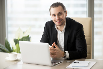 Portrait of handsome friendly smiling businessman in suit in modern office in front of laptop, looking at camera. Positive business person during important meeting with successful negotiation results.