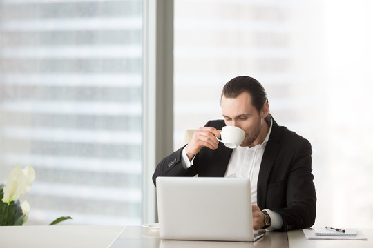 Young handsome businessman drinks coffee while working on laptop at the desk in modern office setting. Daily workflow for businessman or manager concept. Panoramic view. Space for text on the left.
