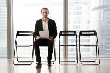 Ambitious smiling young man holds resume or positive business project result, sitting in waiting room. Candidate hopes he will get hired, entrepreneur confident his business plan is successful.