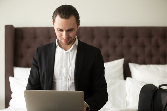 Young smiling businessman working on laptop in bedroom. Freelance entrepreneur remotely works from home, in hotel room while traveling. Business traveler finalizing work project, browsing web concept.