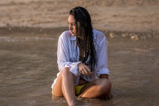 Chica joven con camisa blanca sentada en la playa moj&aacute;ndose con las olas del mar al atardecer.