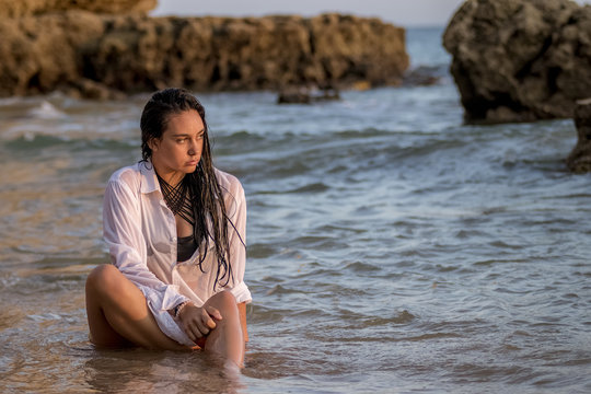 Chica joven con camisa blanca sentada en la playa moj&aacute;ndose con las olas del mar al atardecer.