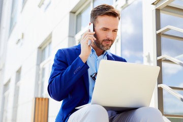 Portrait of focused handsome businessman smiling and talking on phone while working laptop sitting...