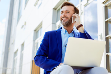 Handsome middle-aged business executive talking on phone smiling and working on laptop while...