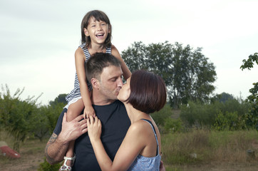 Beautiful happy couple is kissing in nature with their daughter in his shoulders