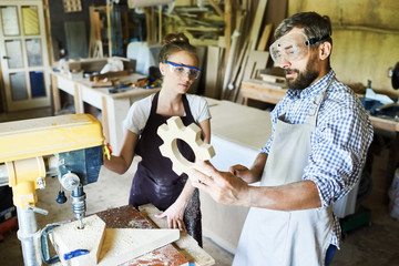 Group of hard-working carpenters looking at just finished wooden detail with concentration while standing at drill press machine, their aprons covered with sawdust