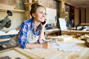 Cheerful young carpenter discussing order details with her customer on smartphone and taking necessary notes, interior of spacious workshop on background - obrazy, fototapety, plakaty