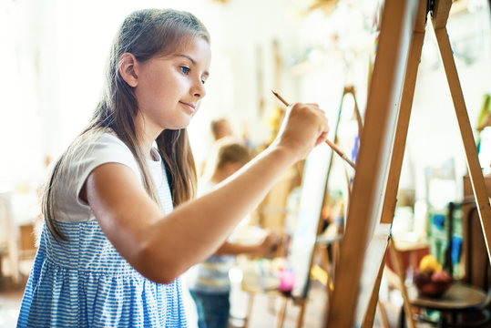 Profile view of pretty little girl wearing striped dress embodying her ideas on canvas with help of paintbrush and watercolors, interior of art studio on background