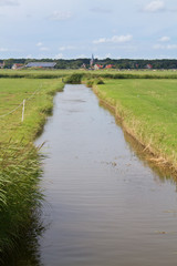 Dutch landscape with a broad ditch between meadows, in the distance a church tower and red tiled roofs of farmhouses