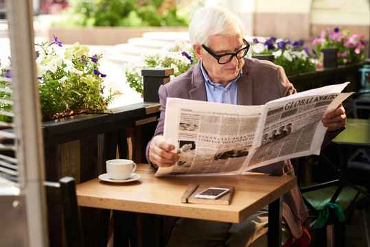Portrait of senior man wearing glasses reading morning newspaper in cafe outdoors