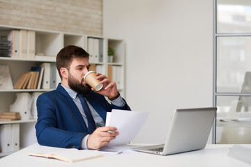 Portrait of successful bearded businessman drinking coffee from paper cup while working at desk in office