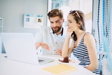 Young couple discussing things looking at laptop while having cake in stylish bright cafe