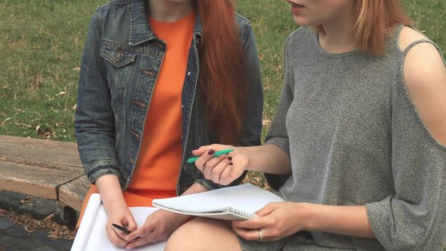 Students sitting on a bench with notebooks. Blonde explaining something. Accent on their hands and notebooks. They are in a park