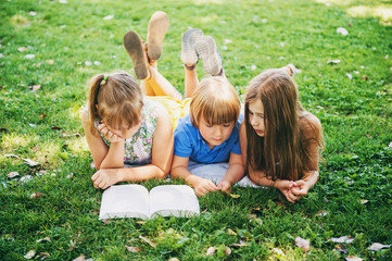 Fototapeta premium Group of three kids lying on green grass and reading story book together. Education for children