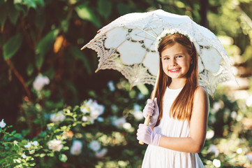 Outdoor portrait of romantic little girl, wearing white dress, gloves, flower headband, holding lace umbrella