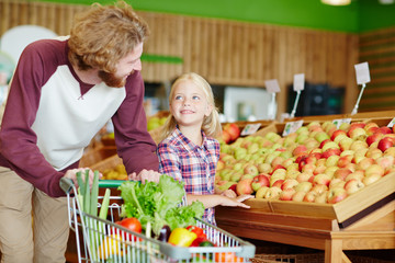 Young man with shopping-cart and his daughter choosing goods in supermarket