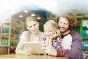 Young parents and little daughter watching curious video in tablet at leisure
