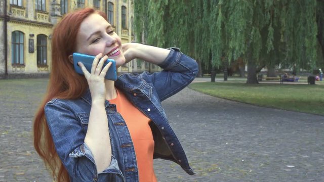 Redhaired girl ralking on phone in front of university. She smiling and laughing. She enjoys herself in her free time