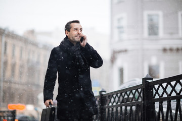 Portrait of handsome bearded entrepreneur with briefcase walking around winter city while talking to his colleague on smartphone, blurred background