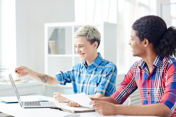 Happy young creative female pointing at laptop display and showing something curious to her colleague