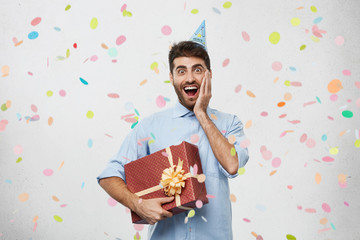 Portrait of excited unshaven birthday guy in formal shirt feeling happy and fascinated about unexpected surprise party thrown by his colleagues, standing in office, holding box in wrapping paper