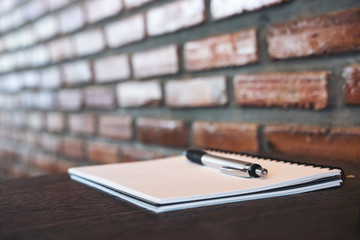 A blank white notebook and silver color pen on wooden table with brick wall background