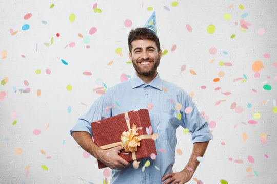 People, fun and celebration concept. Picture of handsome happy young bearded businessman wearing cone hat celebrating his anniversary with colleagues, grinning broadly, showing perfect white teeth
