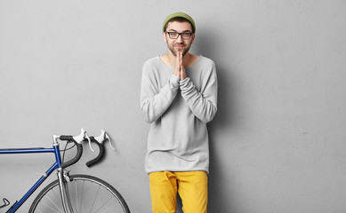 Handsome guy with beard, wearing hat, loose sweater and yellow trousers, keeping hands together having begging look, asking his friend to have ride on bicycles, isolated over grey background