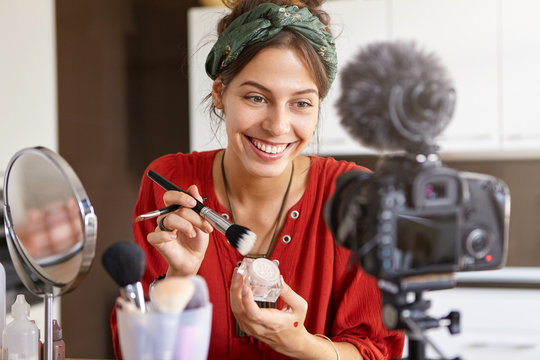 Cheerful teenage girl recording video tutorial at home, looking at camera fixed on tripod, sitting at dressing table, holding brush and powder. Blogging, internet, technlogies and networking concept