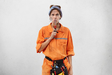 Angry female builder in workwear, asking her companions to be quiet, showing silence sign with screwdriver, being annoyed after busy working day. Untidy female manual wokrer showing hush sign