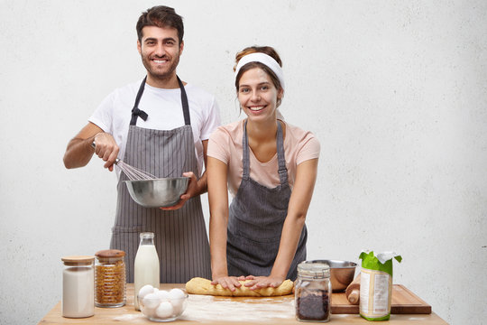 Busy happy family couple make pasrty to bake pie: young beautiful woman rolls dough and husbnad mixes all necessary products for cream in bowl. Team work, cooking, family and culinary concept