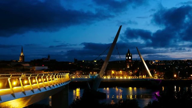 Derry, Ireland. Illuminated Peace bridge in Derry Londonderry in Northern Ireland with city center at the background. Night cloudy sky, reflection in the river.