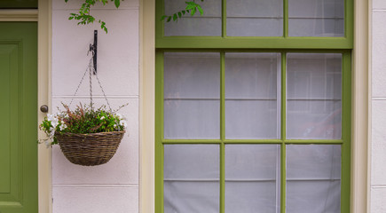 a house facade with a plant pot hanging against a white wall with a green window frame and a green door to the left.