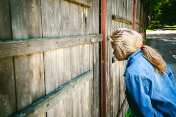 young woman in jeans clothes outdoors. a portrait of a girl with freckles on her face, peeps through a hole in a wooden fence, on a sunny summer day.