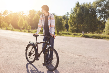 Hipster guy wearing shirt and jeans, standing with his bicycle, looking aside while waiting for his friend to come. Teenager going to school by bike. Handsome stylish boy cyclist. Healthy lifestyle