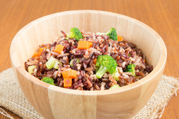 Vegetarian fried rice dish of red brown jasmine rice and riceberry with diced fresh broccoli and carrot in wooden bowl over natural sackcloth on wood table, healthy dish concept (close-up view)