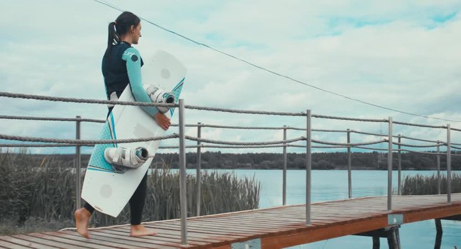 TRACKING young female in a swimsuit walking towards water with a wakeboard in hands, cloudy sky. 4K UHD 60 FPS 