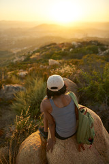 Fototapeta na wymiar Rear view of woman hiker sitting on rock on top of hill while looking at sunset over San Diego California