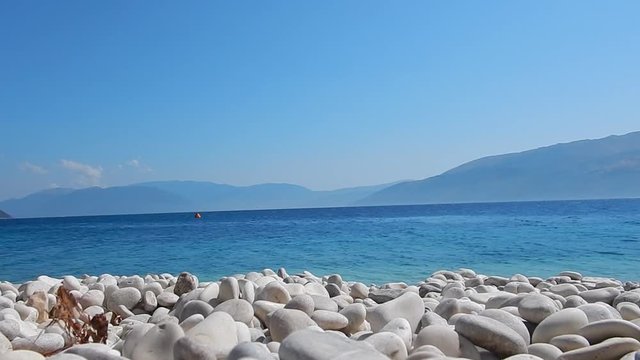 The transparent and turquoise sea in Saint John a beach of white stones, with blue sky and sun, in Ithaca island in Greece and Kefalonia in the background.