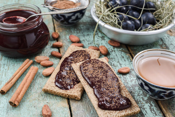Healthy vegan snack of rye crackers with homemade jam served with raw cocoa beans, tea, cinnamon and fresh plums in hay on shabby green wooden table