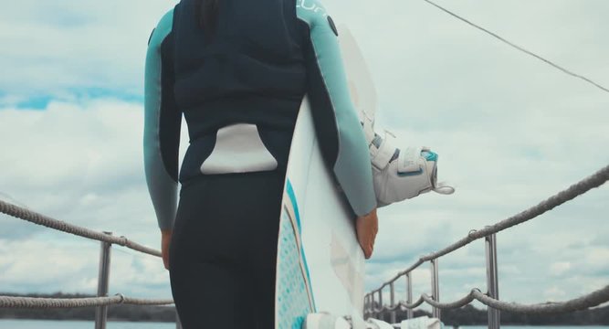 TRACKING back view young female in a swimsuit walking towards water with a wakeboard in hands, cloudy sky. 4K UHD 60 FPS 