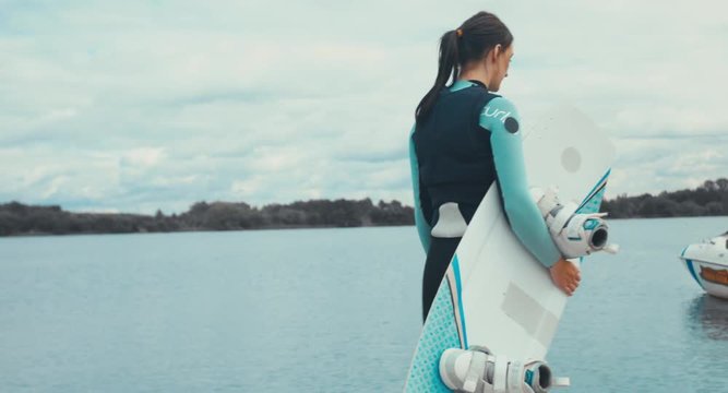 TRACKING back view young female in a swimsuit walking towards water with a wakeboard in hands, cloudy sky. 4K UHD 60 FPS