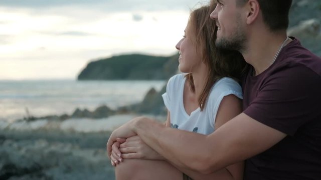 Man woman sitting in hug on shore on stone watching sunset